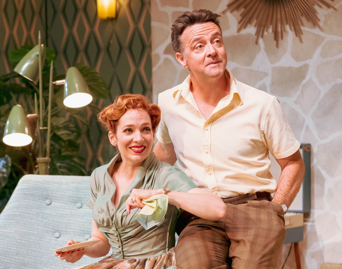 Judy (Katherine Parkinson), dressed in 1950s clothing, sits happily in a lounge holding a cloth and a plate. Her husband sits next to her