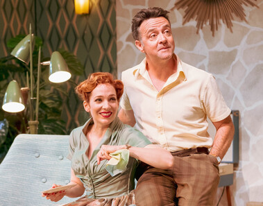 Judy (Katherine Parkinson), dressed in 1950s clothing, sits happily in a lounge holding a cloth and a plate. Her husband sits next to her