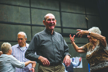 A group of elderly people dance together in the theatre, laughing and smiling