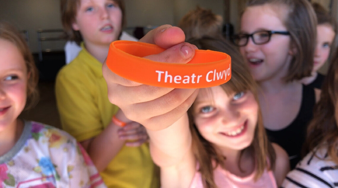 A group of children stand together. In the centre a girl holds up an orange wristband which has the text ‘Theatr Clwyd’ on it.