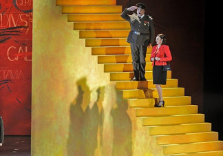 A man and a woman stand on some steps, the man dressed in military clothes while saluting, the women wearing casual clothes. By the stairs, several men look onward also dressed in military attire