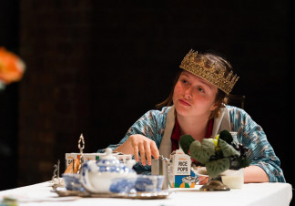A woman sits at a breakfast table wearing a cloak and a crown