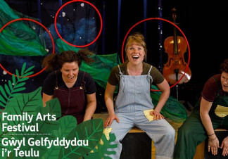 Three people sit cheerfully together, one playing percussion on a box. In the background is a cello, leaves and a starry sky
