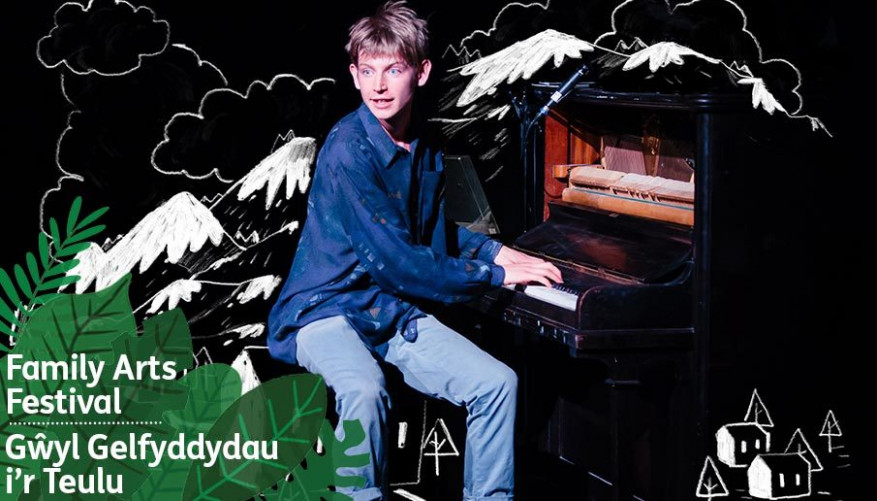 A boy sits and plays a piano in front of an illustrated background of mountains in black and white