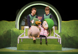 Two puppeteers control a large puppet of a duck, and a large puppet of a young girl, which are sat on a bench in front of a green background