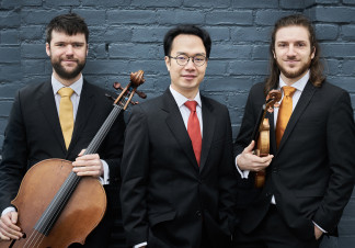 The members of the trio stand together in suits. The one on the left is holding a cello, while the leftmost one is holding a violin