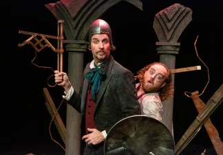 Two men in 19th century clothing stand together. One is dressed in a suit, wearing a medieval helmet and a holding a metal instrument. The man on the left is holding a shield and a wooden plank. They are stood in front of ruined pillars
