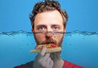 A ginger man with a bushy beard bites on a slice of pizza. His head submerged up to the bridge of his nose under water